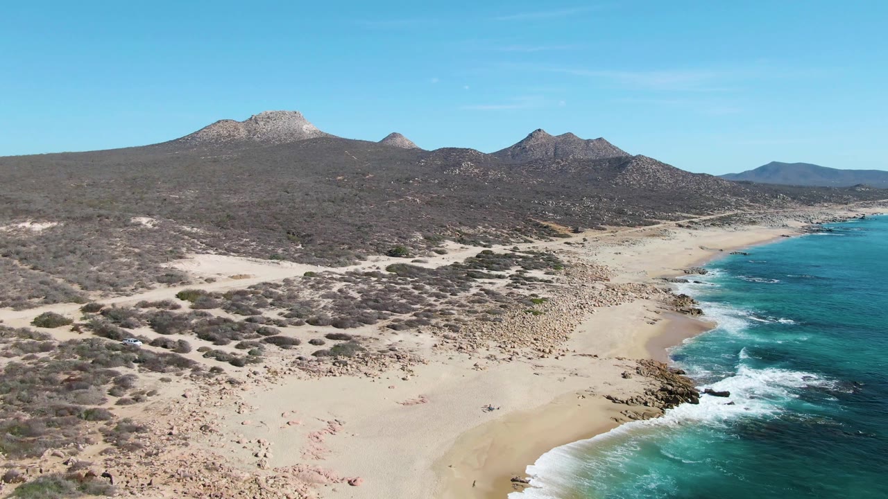 Aerial frame at Buzzards Beach, Baja California Sur, showing a sandy shoreline curving beside turquoise water with desert scrub in the foreground and rocky mountains beyond.