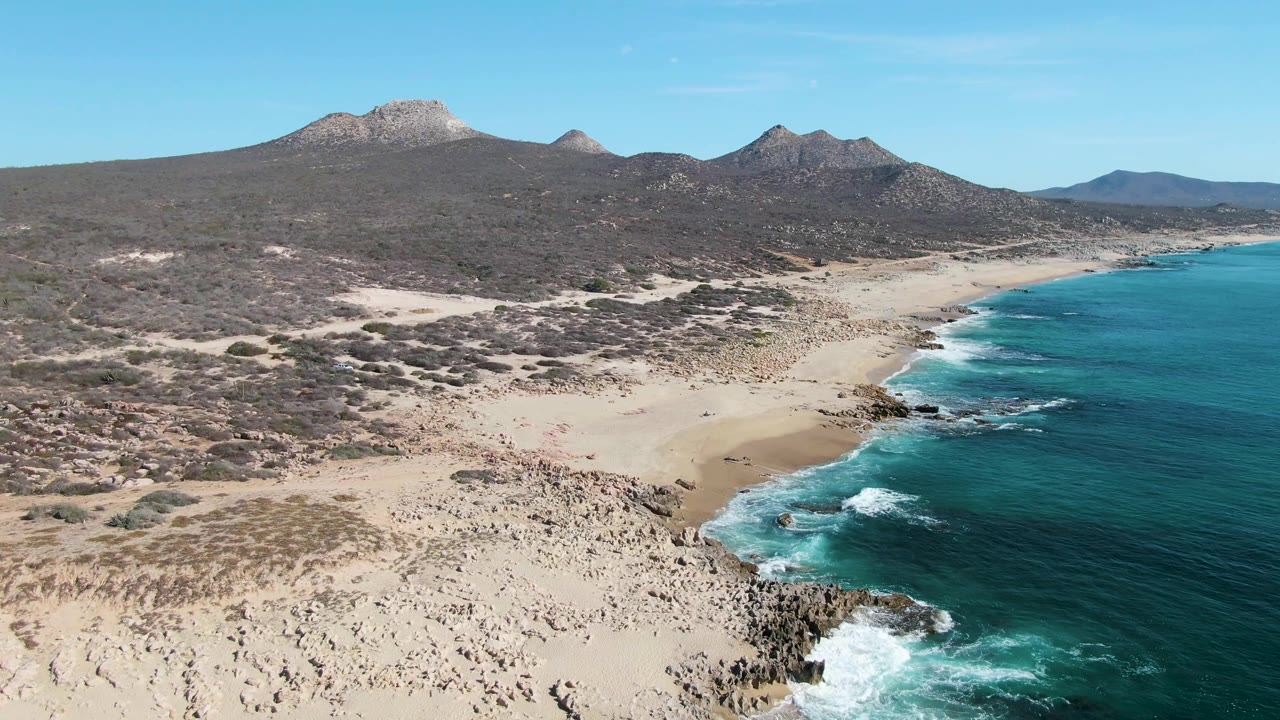 Aerial frame over Buzzards Beach, Baja California Sur, showing pale sand, rocky shoreline, turquoise water to the right, arid scrubland, distant mountains, and no visible people.