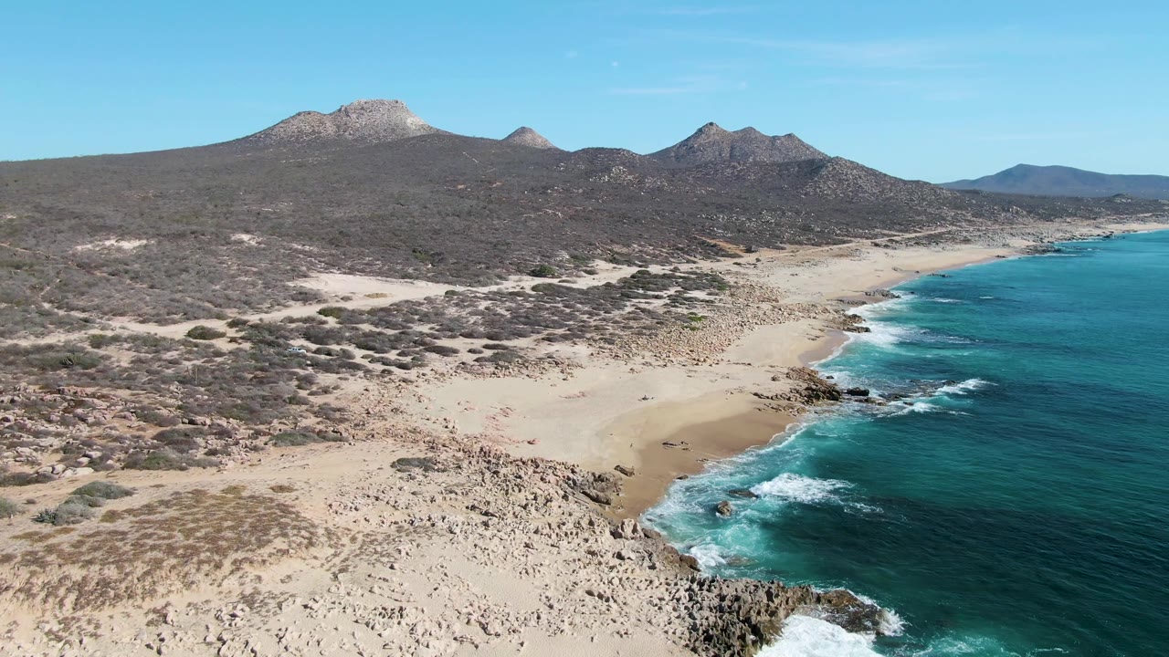 Aerial frame over Buzzards Beach with white surf curling against rocks, a broad sandy cove, dry scrub terrain behind it, mountains on the horizon, and no visible people.