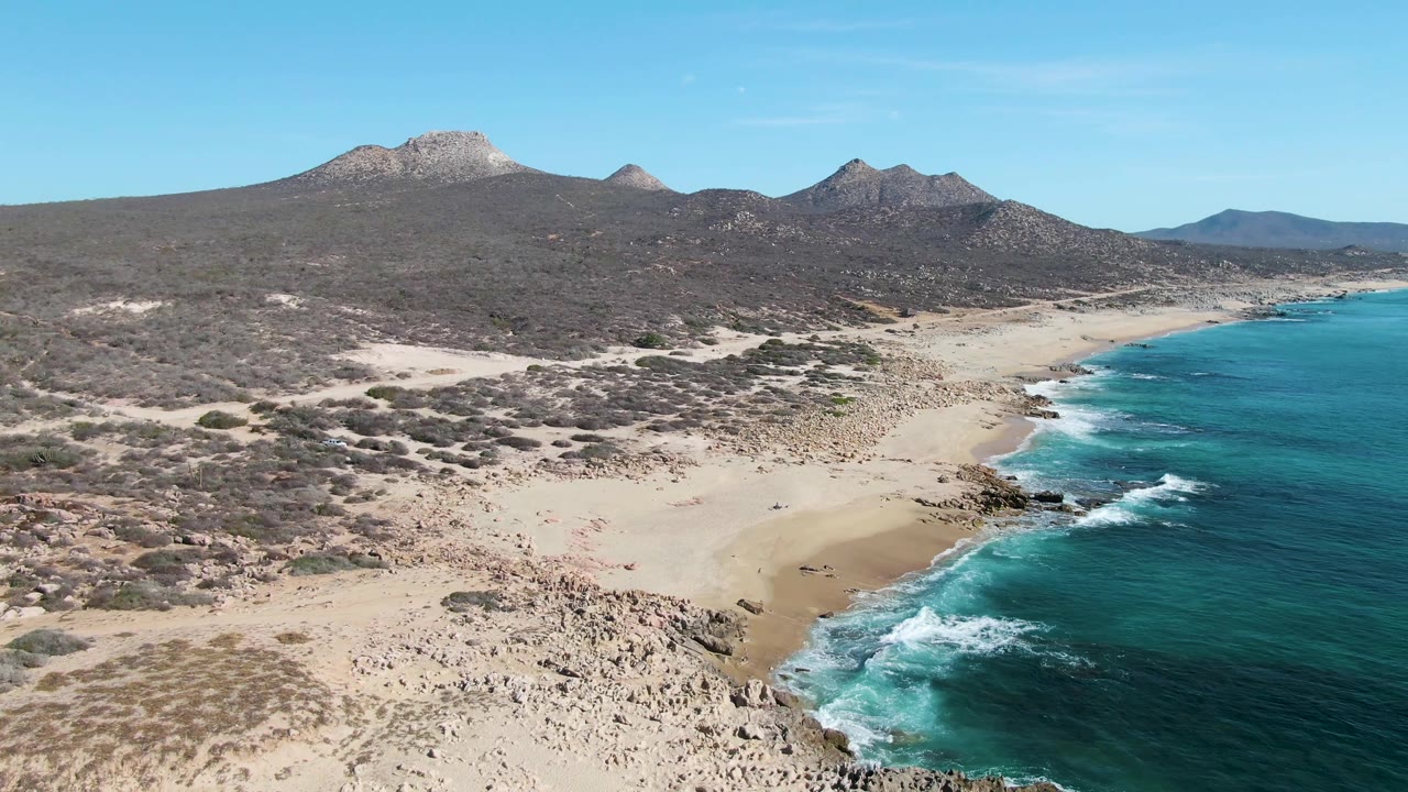 Aerial frame over Buzzards Beach showing a longer stretch of turquoise coastline, pale sand, scattered rocky patches, dry land, distant mountains, and no visible people.