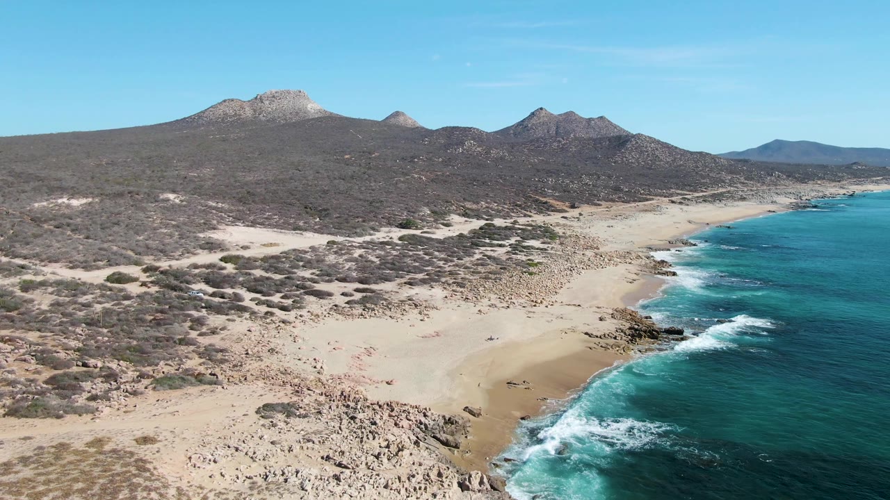 Aerial shoreline frame with rocky foreground, sandy beach, turquoise sea, scrubland, and mountains.