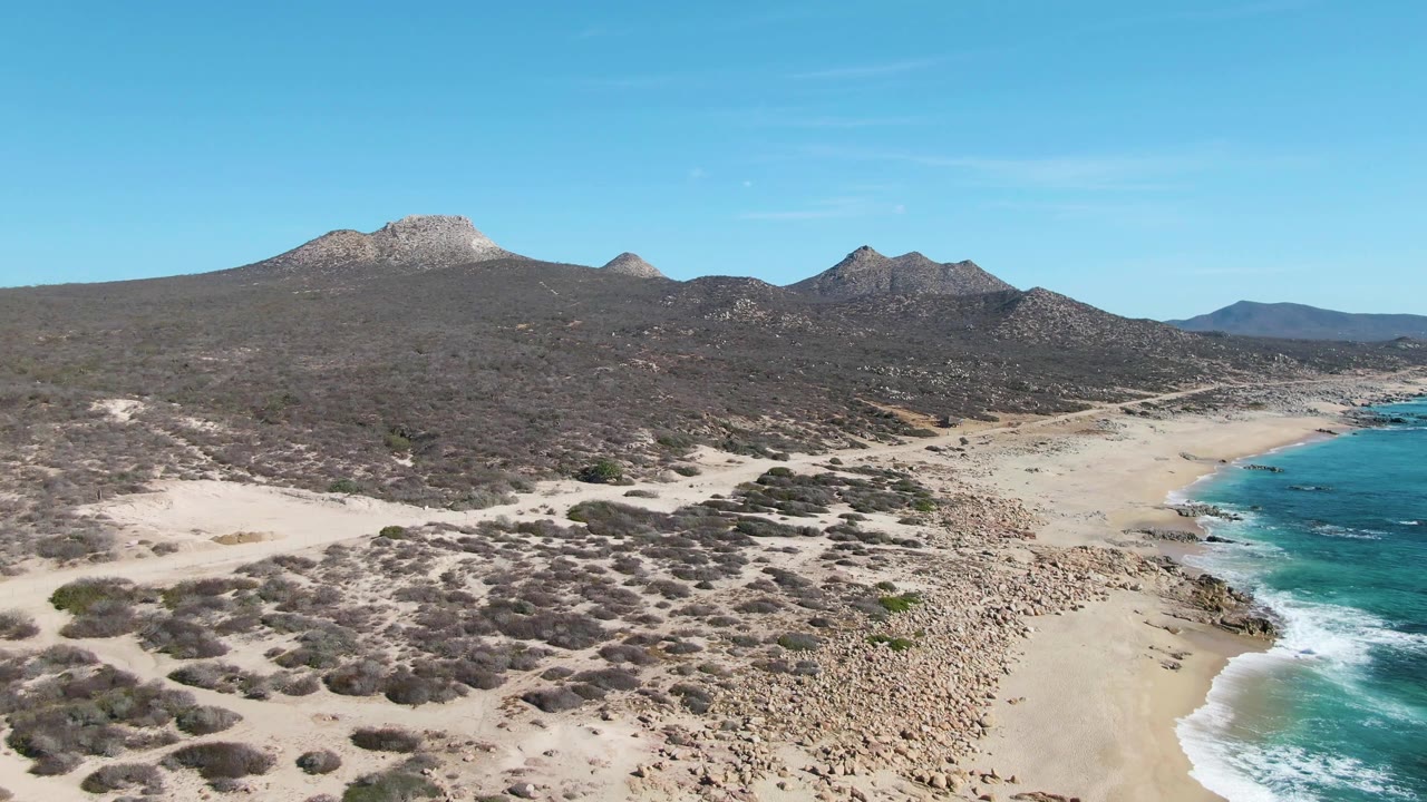 Wide aerial frame over Buzzards Beach in Baja California Sur, with pale dunes and scrub in front, turquoise surf at the right edge, mountains across the background, and no visible people.