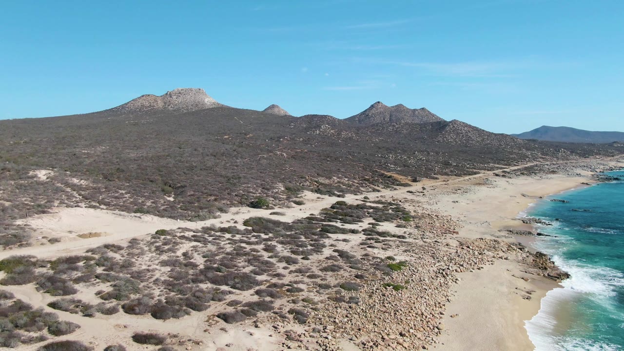 Buzzards Beach aerial view with dense low scrub across sandy foreground, a pale beach and rocky strip below, turquoise water on the right, distant hills and mountains, and no visible people.