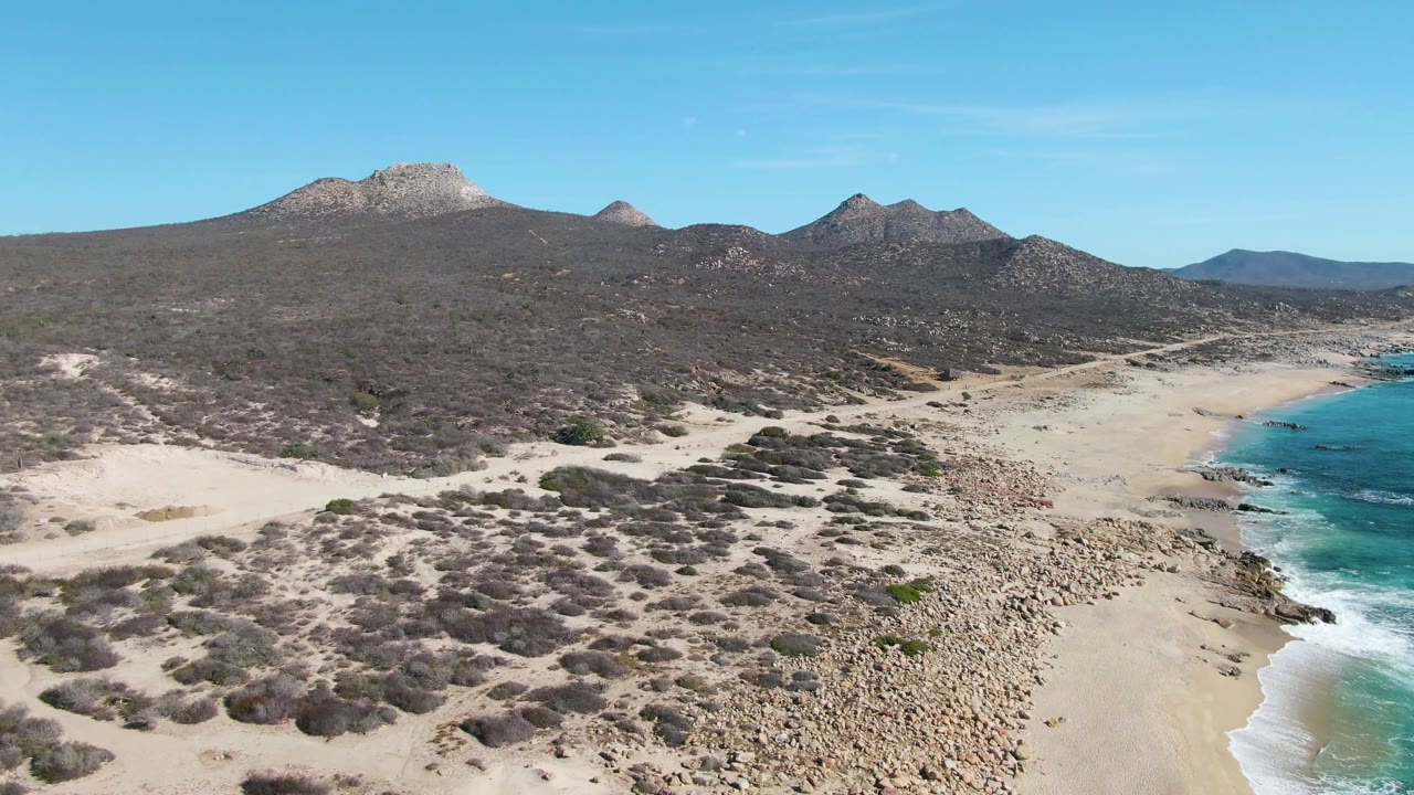 Aerial frame at Buzzards Beach showing a broader sweep of dunes and scrub, the rocky sandy shore leading toward turquoise water at frame right, mountain ridges behind, and no visible people.