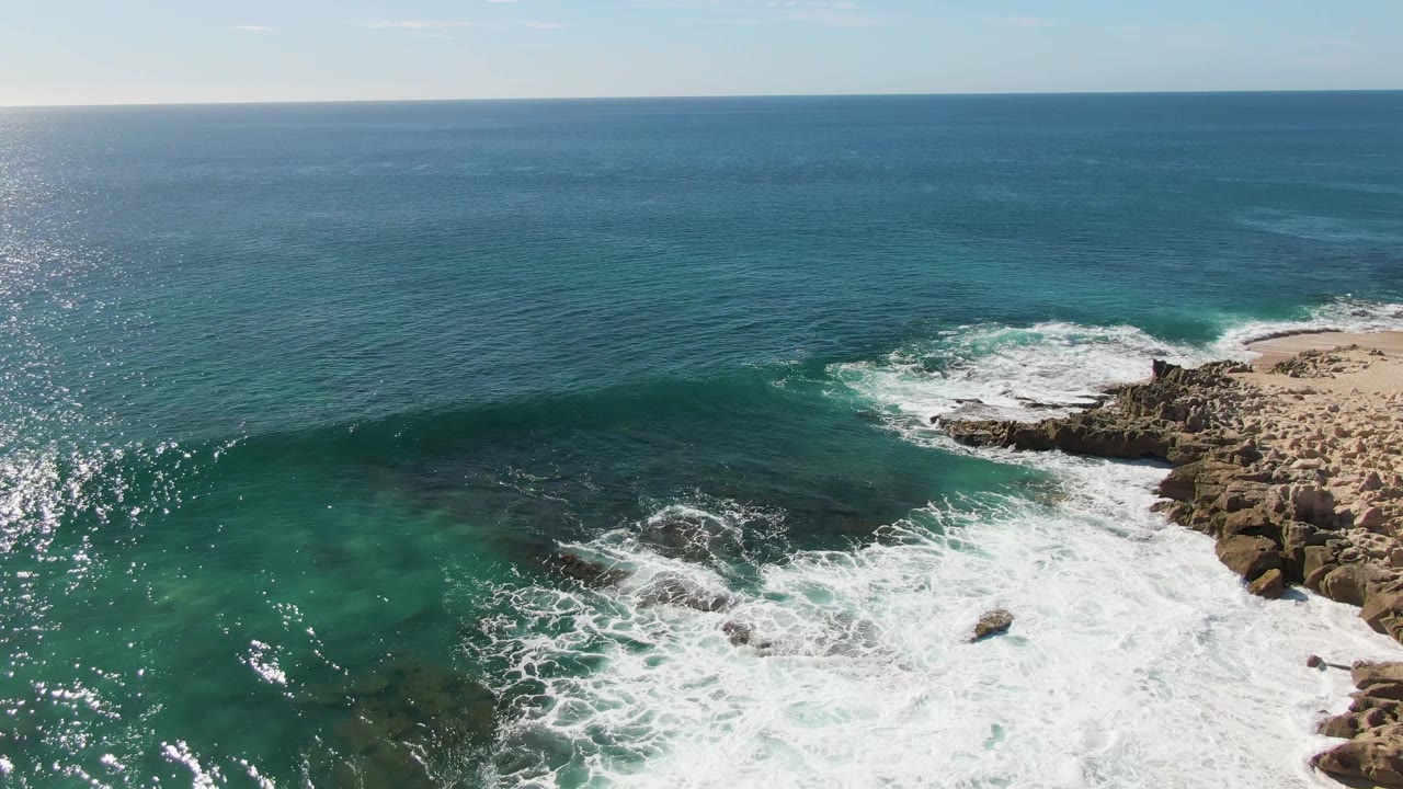 At Buzzards Beach in Baja California Sur, turquoise water rolls into white foam over shoreline rocks with a small sandy beach at right and no visible people.