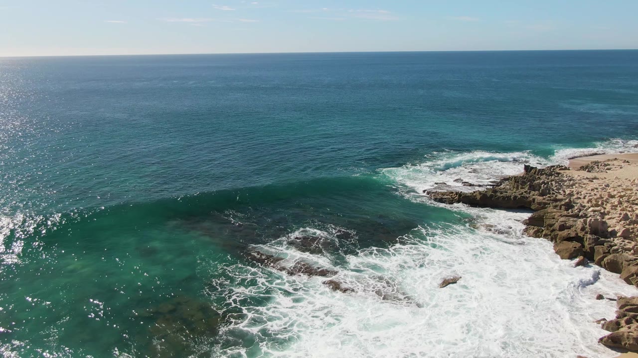 At Buzzards Beach in Baja California Sur, a larger sheet of white foam spreads across the rocks below the blue ocean horizon, with no visible people.