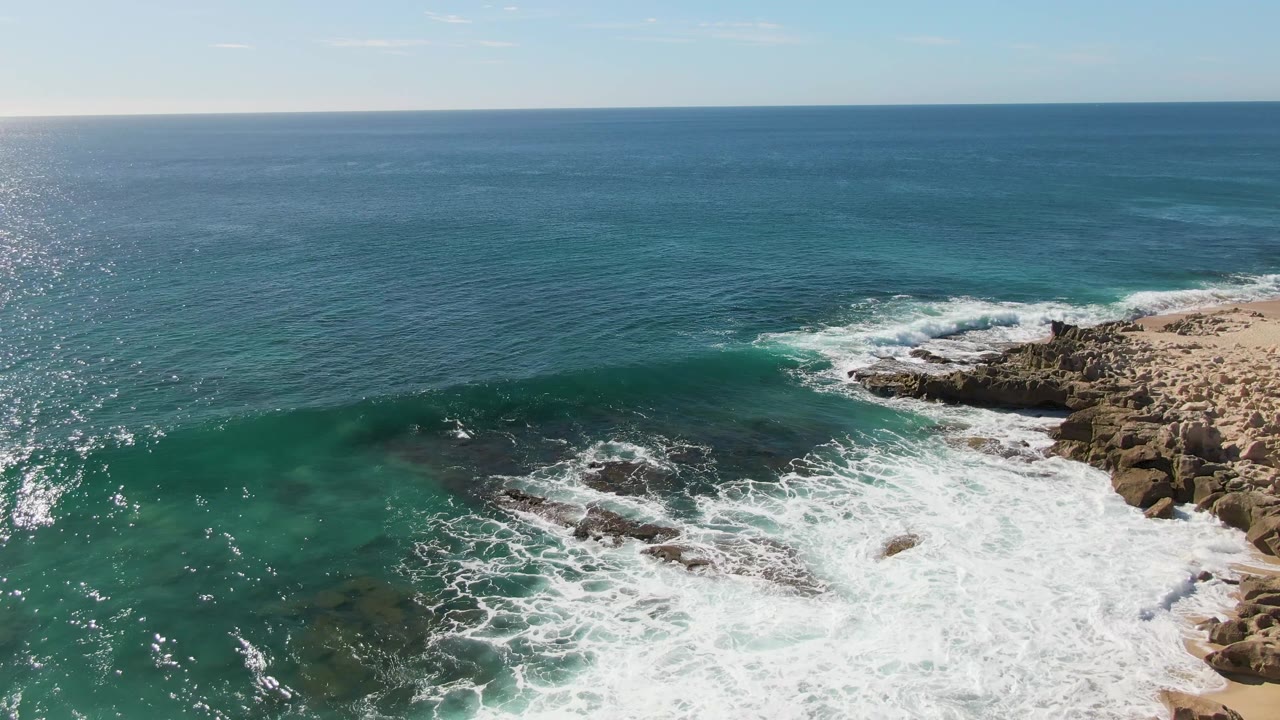 At Buzzards Beach in Baja California Sur, a green-blue wave breaks toward the rocky right edge while foam washes over the lower shoreline and no people are visible.