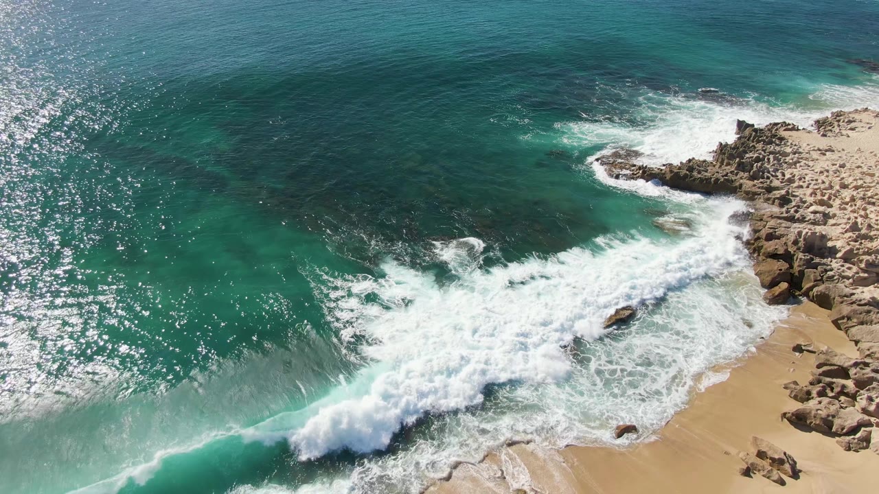 Aerial view of waves breaking onto rocks and sand beside turquoise ocean.