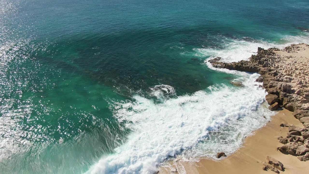 At Buzzards Beach in Baja California Sur, Mexico, white foam expands over the sand and rock edge while darker blue ocean sits beyond the turquoise shallows, with no visible people.