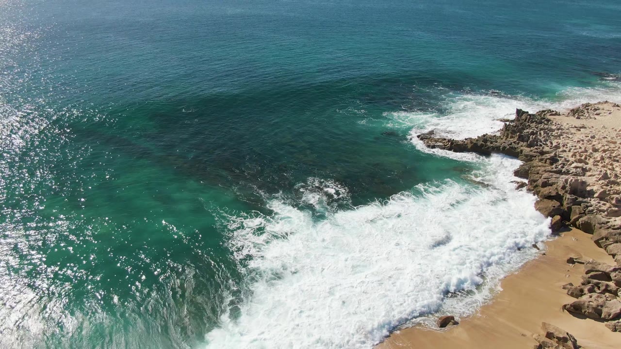 Aerial frame at Buzzards Beach in Baja California Sur, Mexico, showing heavy whitewater across the lower sand, rocky formations on the right, clear blue-green water, and no visible people.