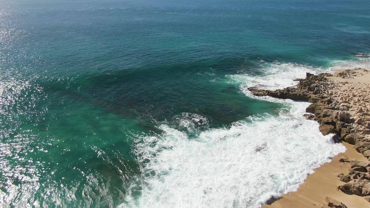 Wide aerial view of Buzzards Beach in Baja California Sur, Mexico, with the horizon visible, calm deep blue water offshore, foamy surf near the rocks, sandy beach at right, and no visible people.