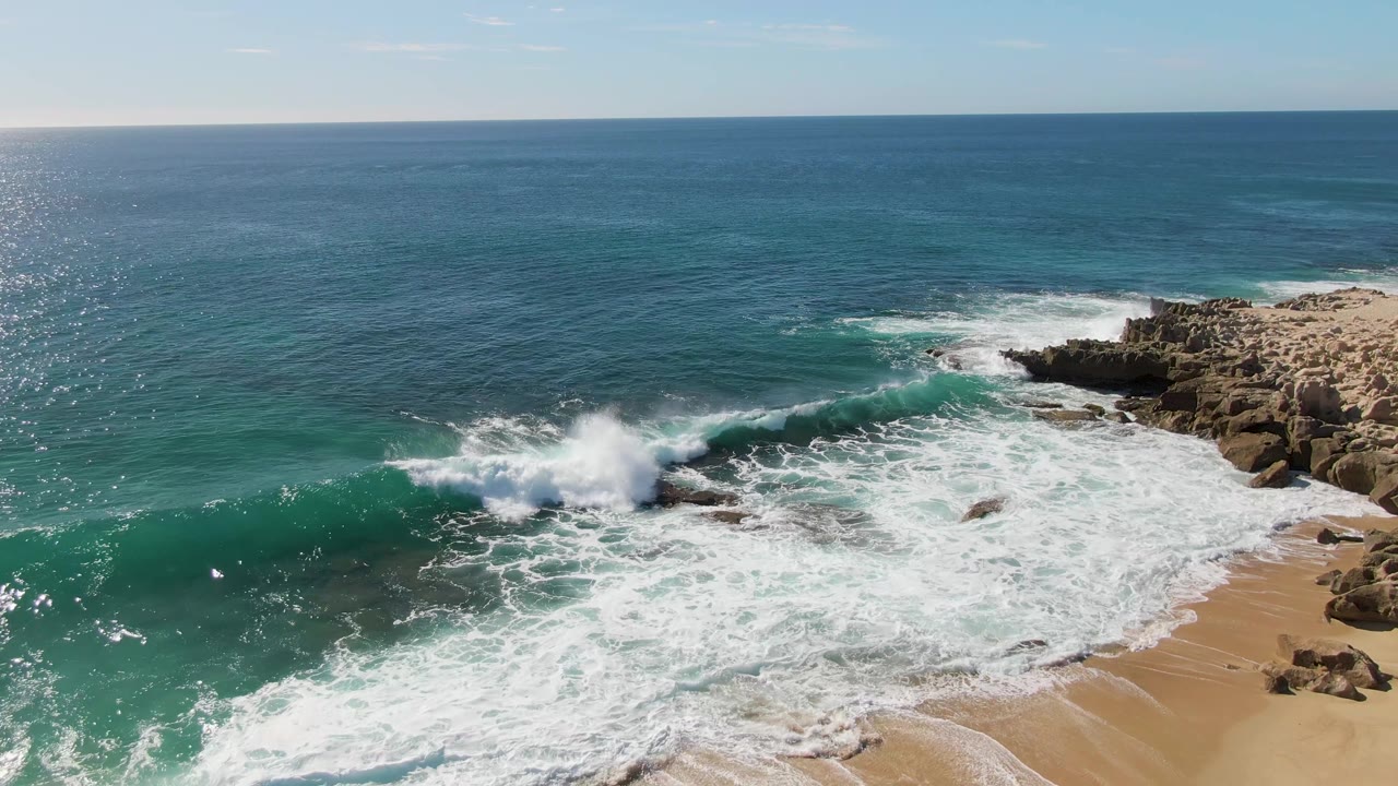 At Buzzards Beach in Baja California Sur, a turquoise wave begins breaking toward rocky outcrops beside tan sand, with bright ocean glare and no visible people.