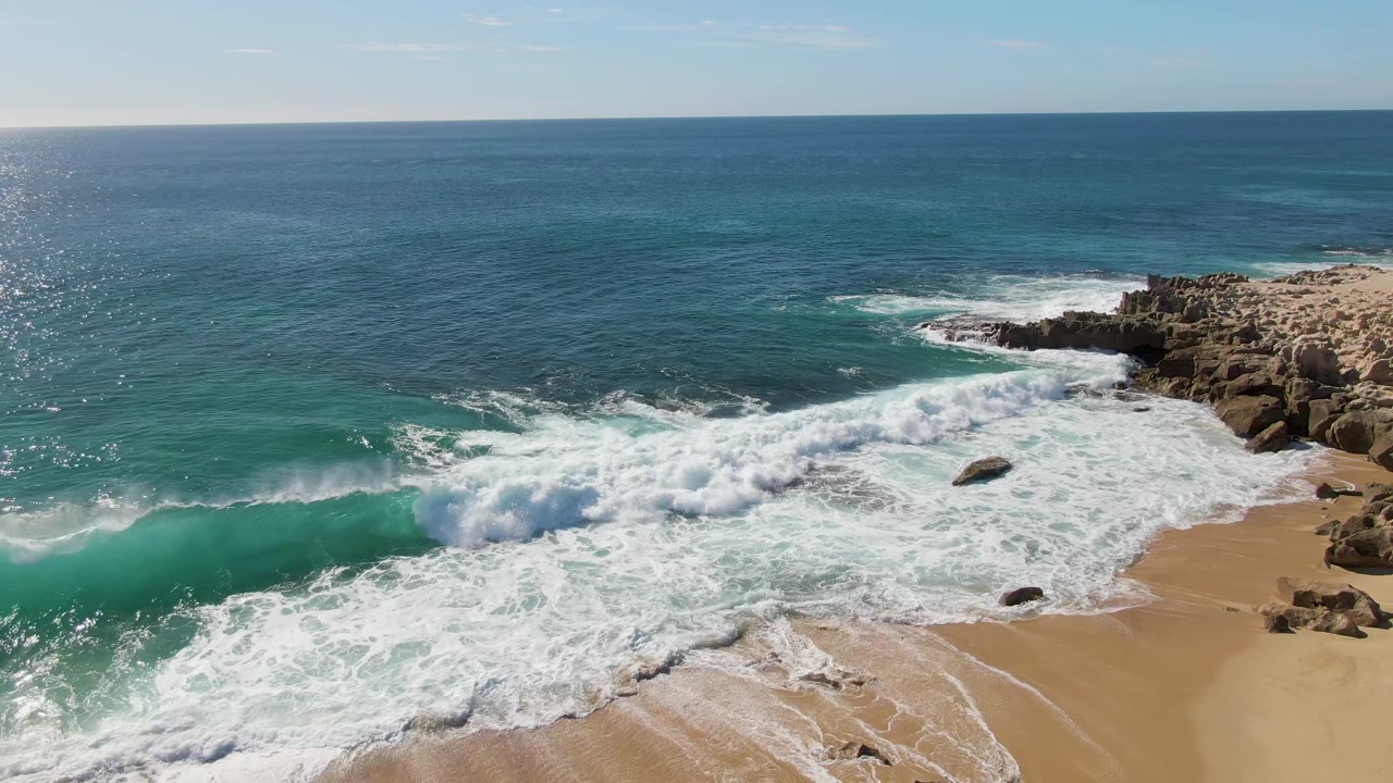 At Buzzards Beach in Baja California Sur, the breaking wave has filled much of the shore with white foam, with dark rocks on the right and open blue water beyond, no visible people.