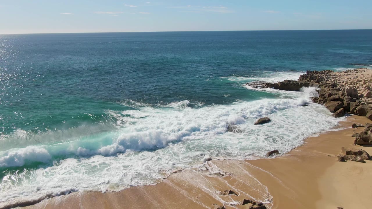 At Buzzards Beach in Baja California Sur, a larger turquoise wall of water rolls across the lower left while foam from the previous break moves over sand and rocks, with no visible people.