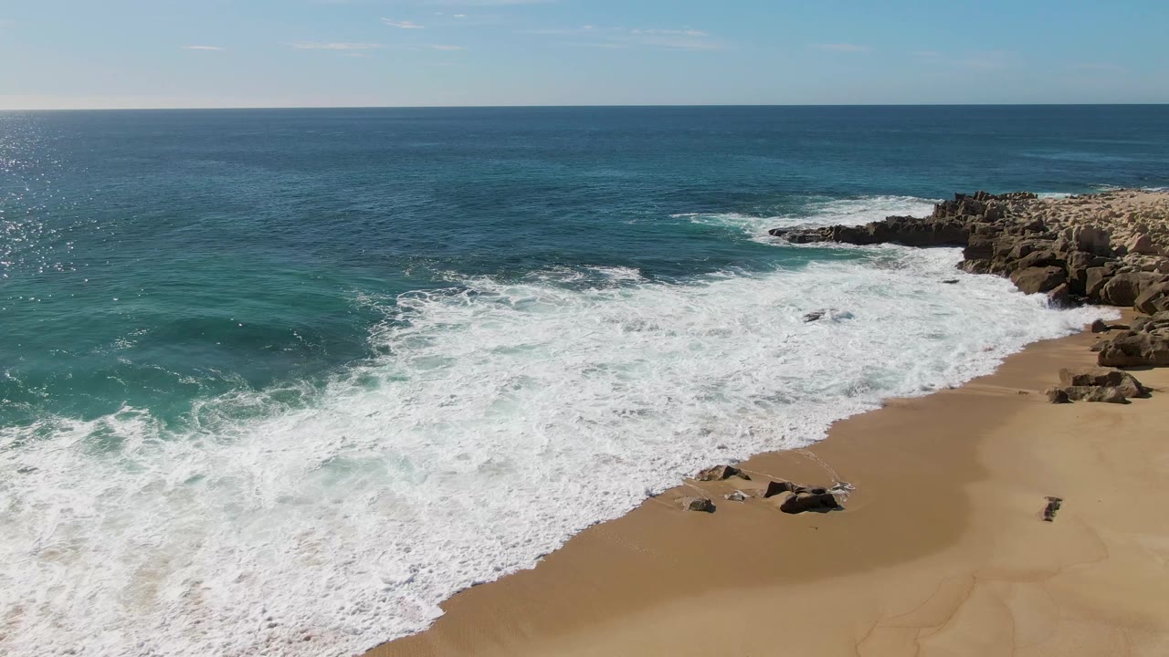 Aerial frame from Buzzards Beach, Baja California Sur, Mexico, with a broad sheet of white foam moving over the sandy shore, darker blue water beyond, rocks at upper right, and no visible people.
