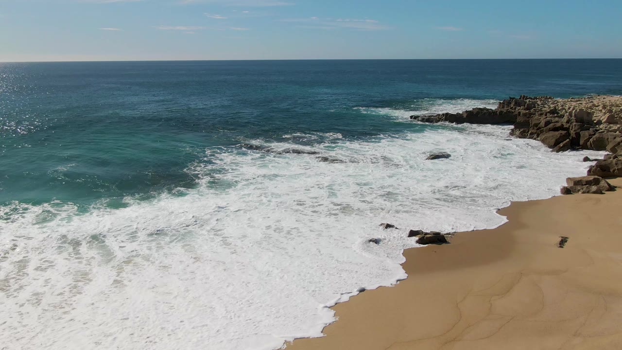 Aerial frame from Buzzards Beach, Baja California Sur, Mexico, showing receding white water across the sand, green-blue surf at left, dark rocks along the right shoreline, and no visible people.