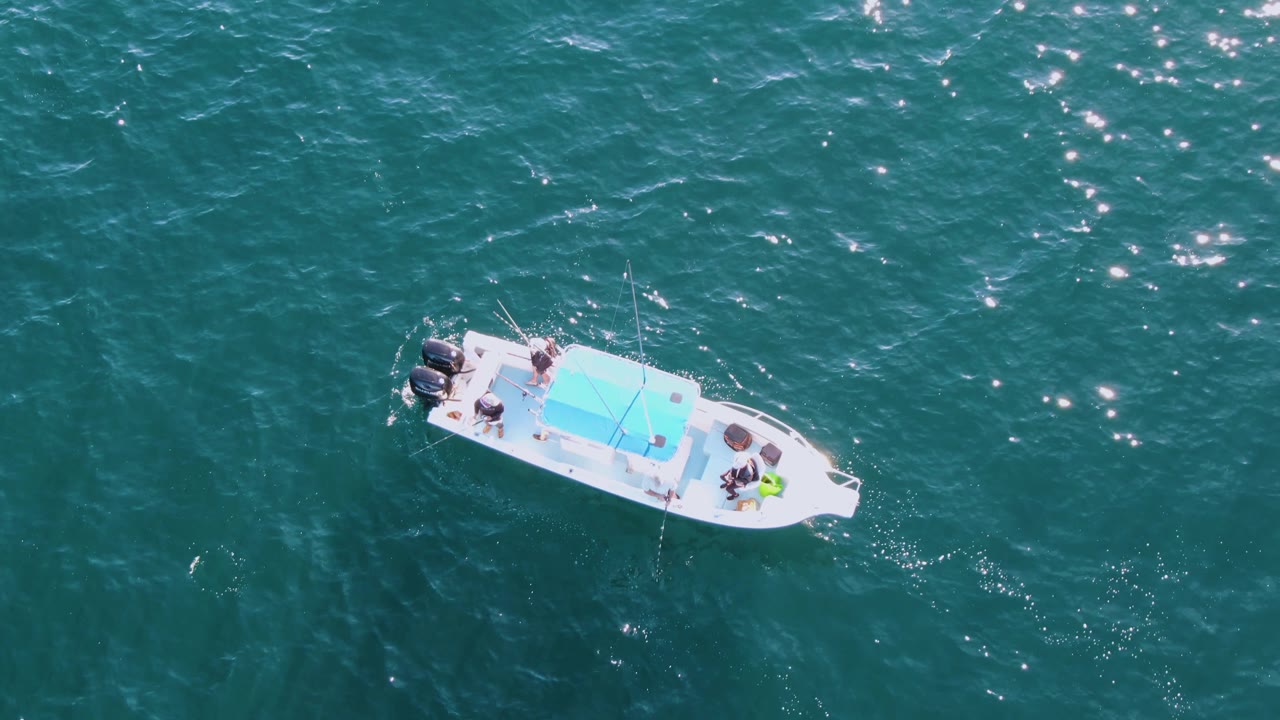 The boat remains isolated on teal ocean water at Buzzards Beach, with fishing rods visible and sun sparkles concentrated across the upper part of the frame.