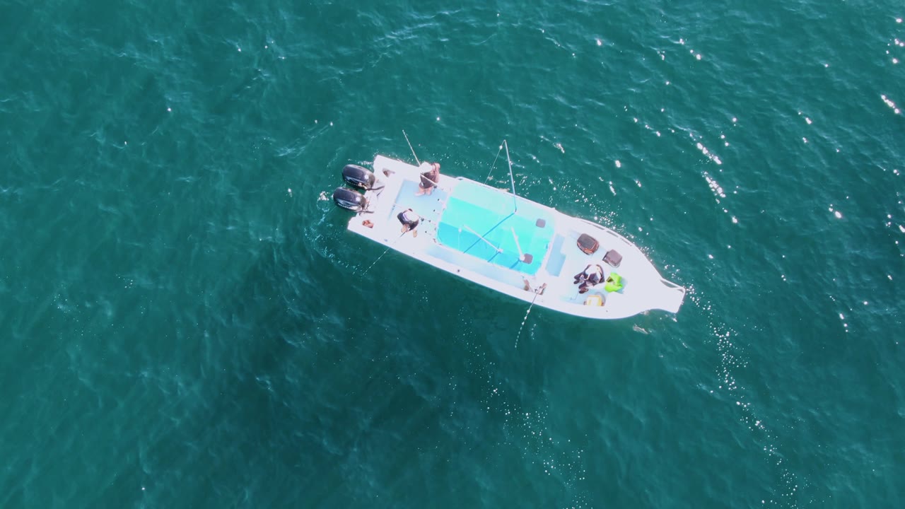 Overhead Buzzards Beach frame with the motorboat shifted across open teal water, its twin outboard motors, blue deck panel, and small groups of people still visible.