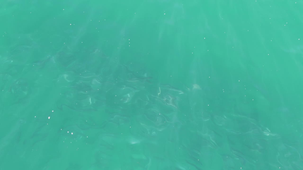 Turquoise ocean surface in Buzzards Beach with darker wavering shapes under the water, tiny sun glints near the middle, and no visible people.