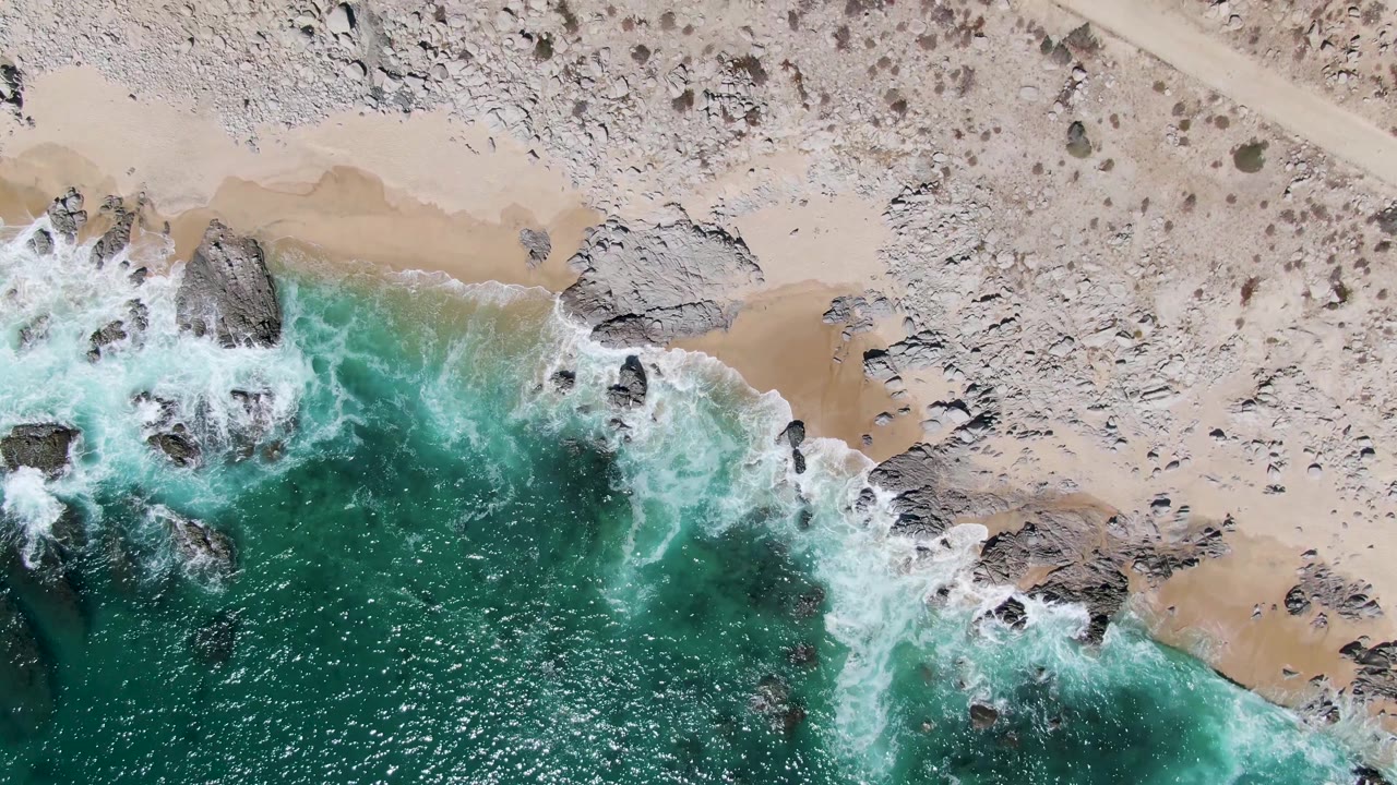 Overhead view of turquoise surf, rocks, sand, desert scrub, and a dirt road.