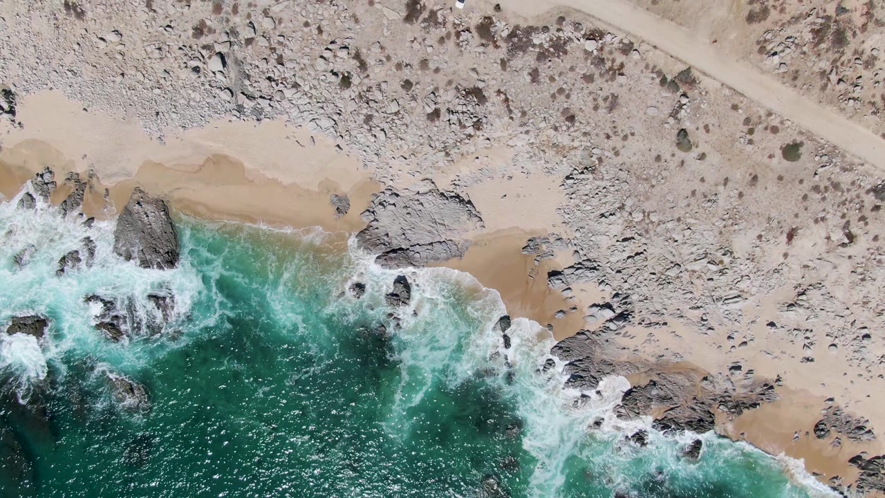 Buzzards Beach aerial view shows white foam spreading around shoreline rocks, tan sand above the water, a diagonal dirt road, a small white car, and no visible people.