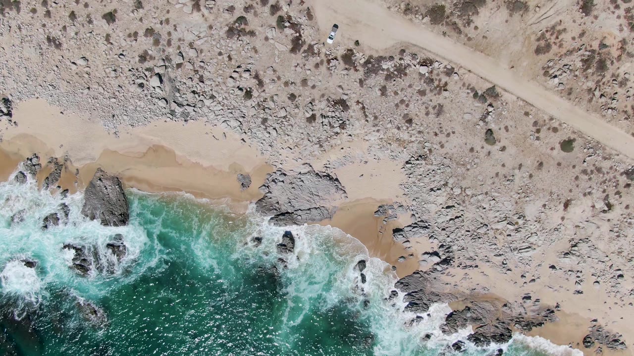 The overhead frame at Buzzards Beach places the white car near the upper road while waves curl around rocks and narrow sand pockets below, with no visible people.