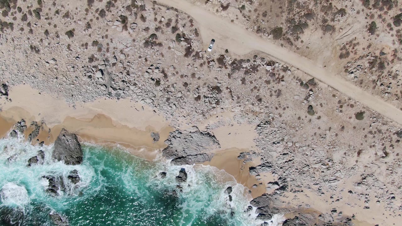 Buzzards Beach appears from above with the dirt road crossing the scrubby upper frame, surf washing over rocks at the lower edge, tan sand between them, and no visible people.