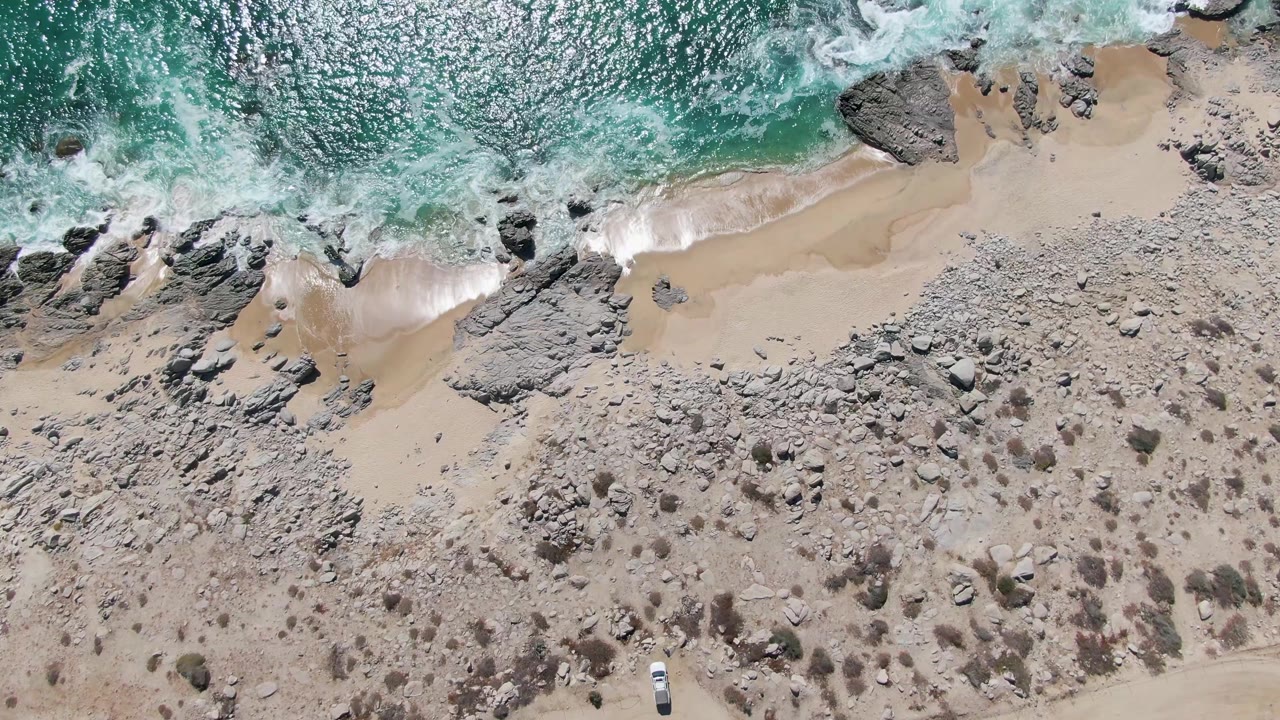 Buzzards Beach aerial frame with white foam pushed closer to the sand, large rocks along the waterline, pale beach below, rocky inland ground, and no visible people.