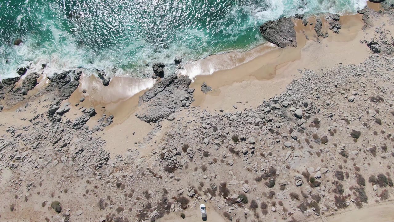 Top-down Buzzards Beach view with bright textured ocean, foamy surf tracing the sand, a broad rocky shelf below, sparse scrub on dry ground, a dirt road, a small car, and no visible people.