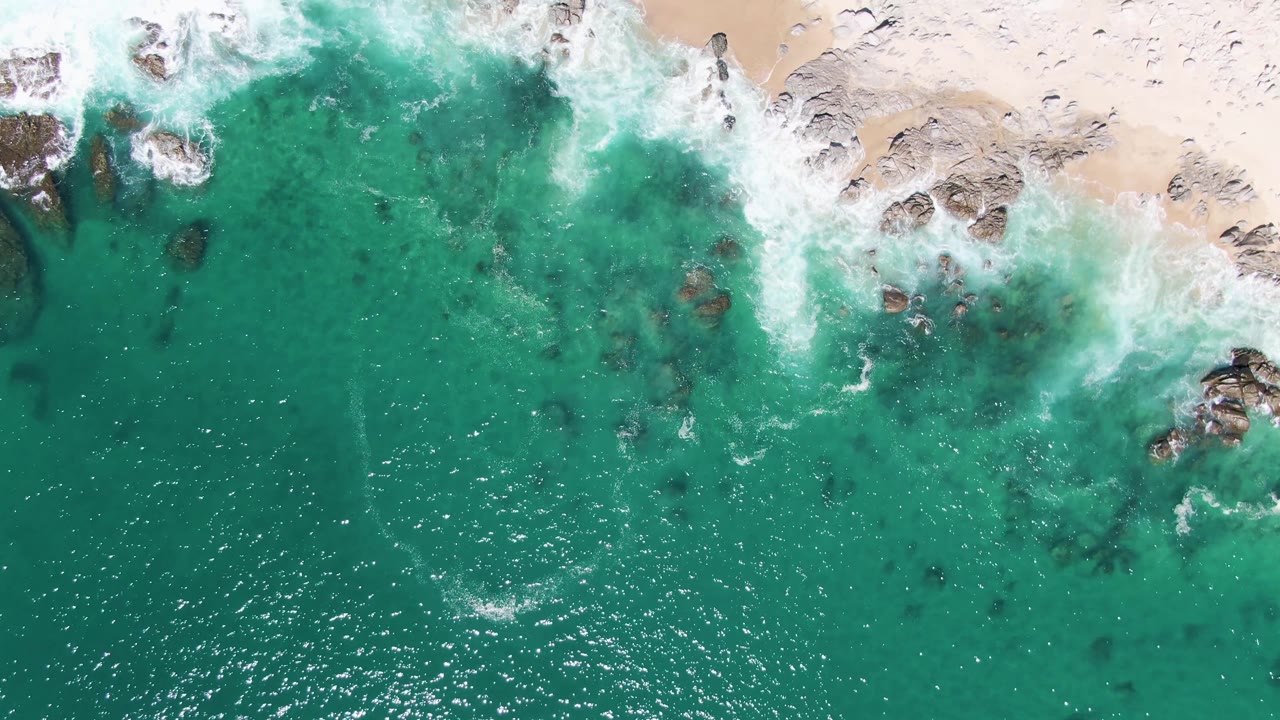 Overhead turquoise surf hitting rocks beside pale sand