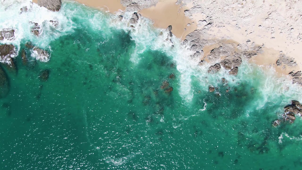 Aerial frame at Buzzards Beach showing white water spreading around dark rocky outcrops in clear turquoise ocean, with no visible people.