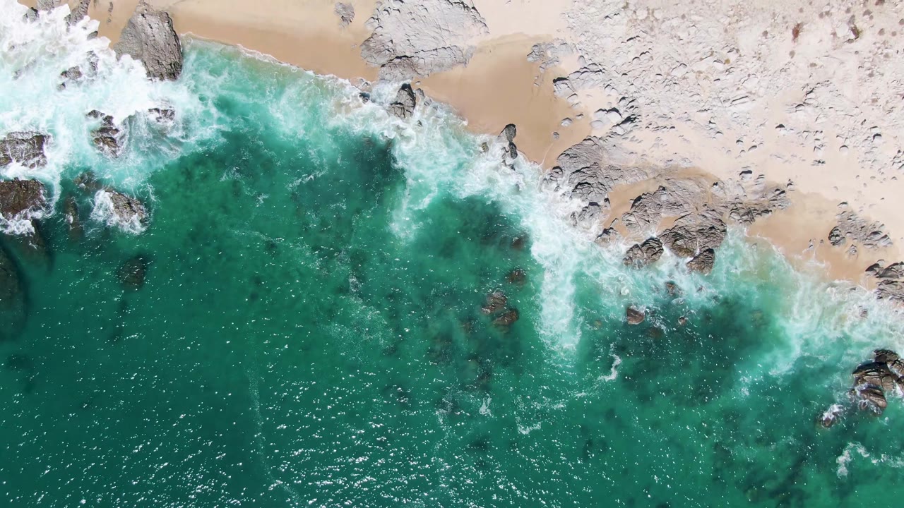 Top-down coastal frame from Buzzards Beach with pale sand across the upper edge, rocks along the surf line, turquoise water below, and no visible people.
