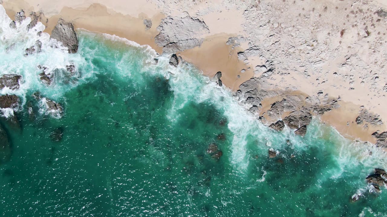 Overhead Buzzards Beach view with a larger band of white surf crossing dark rocks and green-blue water, while the sandy beach remains empty of visible people.