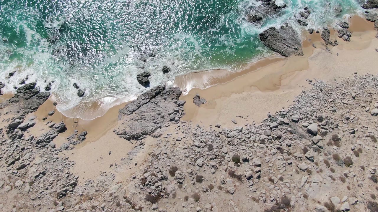 Overhead aerial frame of Buzzards Beach in Baja California Sur with turquoise water, white foam, pale sand, dark rocks, scattered stones, and no visible people.