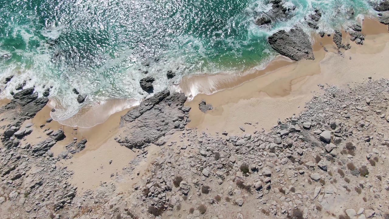 White surf curls along pale sand beside exposed rocks.