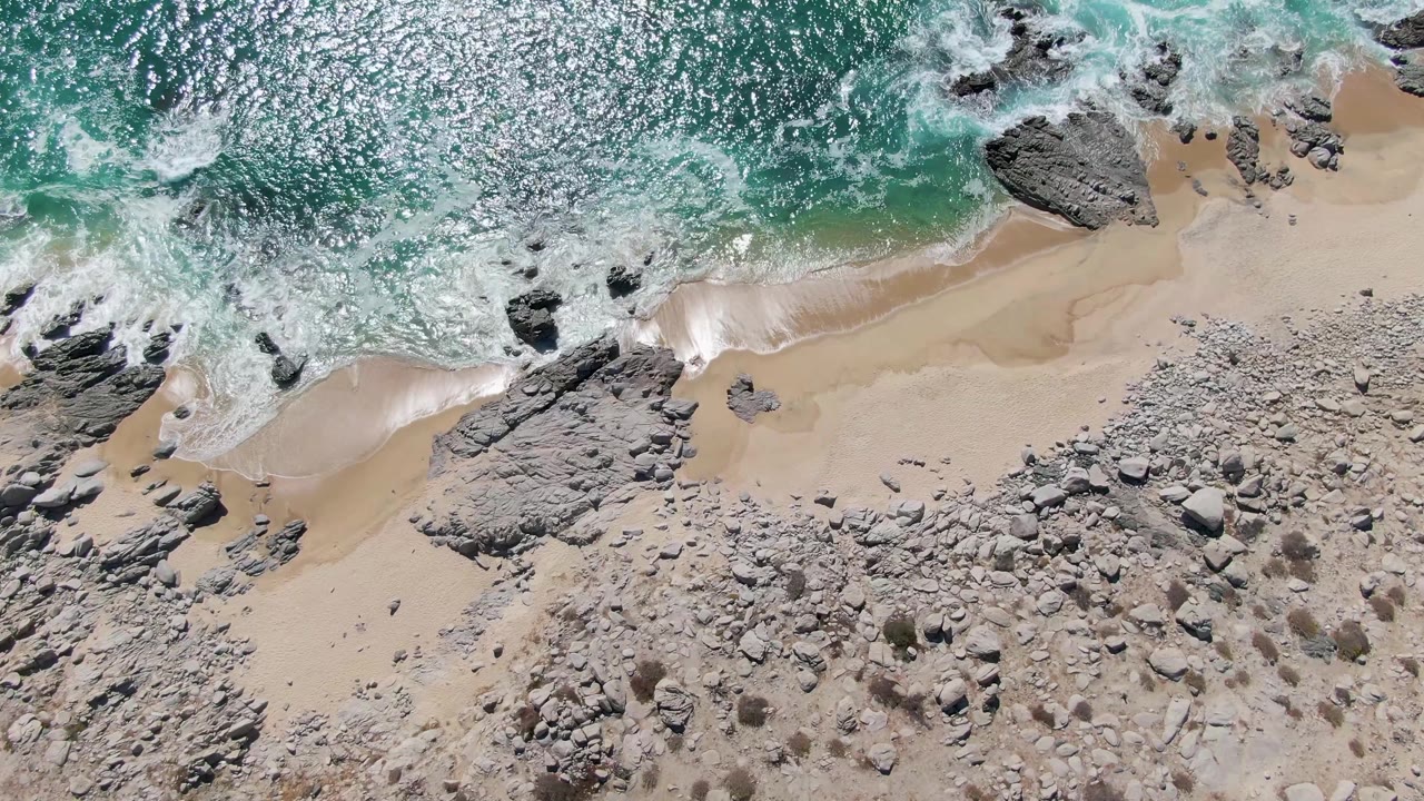 Overhead frame from Buzzards Beach with bright sun glitter across turquoise water, wave foam breaking around dark rocks, a pale sandy strip, scattered stones, and no visible people.