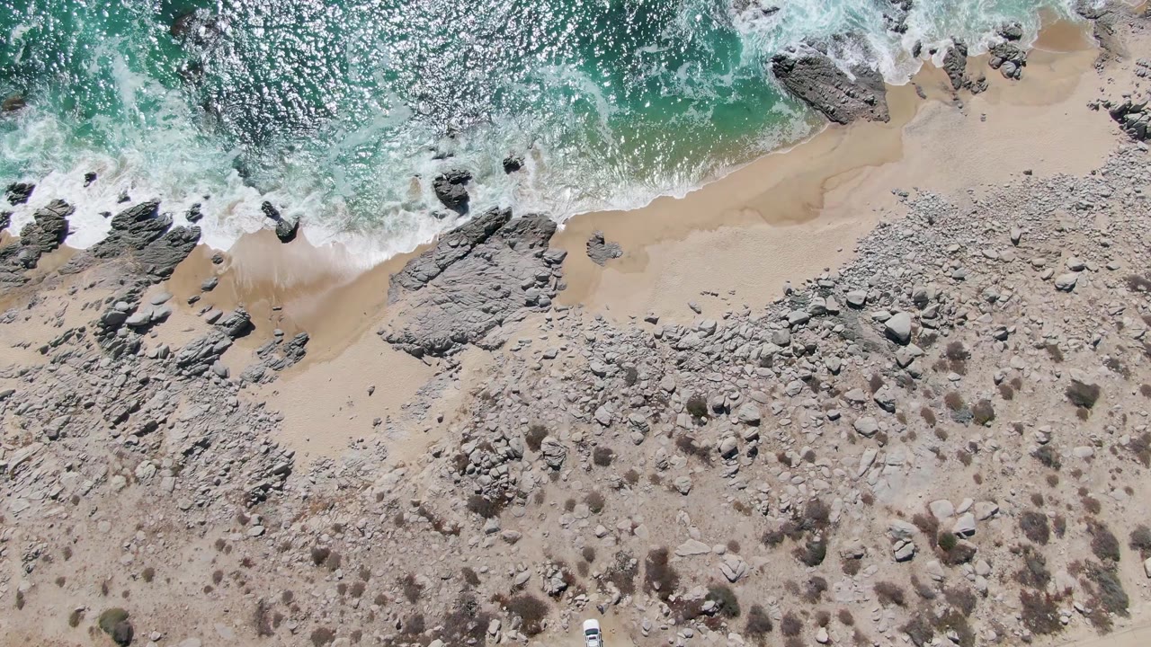 Aerial frame over Buzzards Beach, East Cape, Baja California Sur, showing turquoise surf, white foam, pale sand, rocky terrain, a small white vehicle near the lower edge, and no visible people.