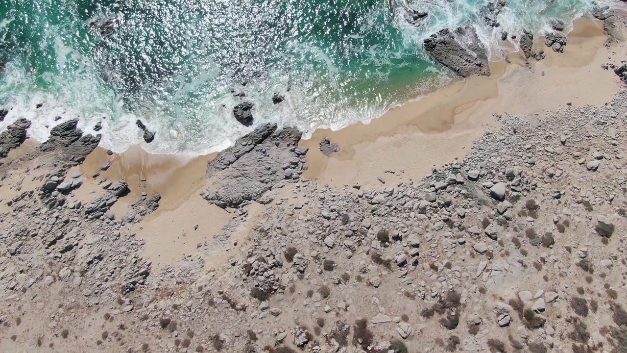 Top-down Buzzards Beach frame with sun glitter on turquoise water, a broken white wave line, narrow sand, clustered shoreline rocks, dry rocky ground, and no visible people.