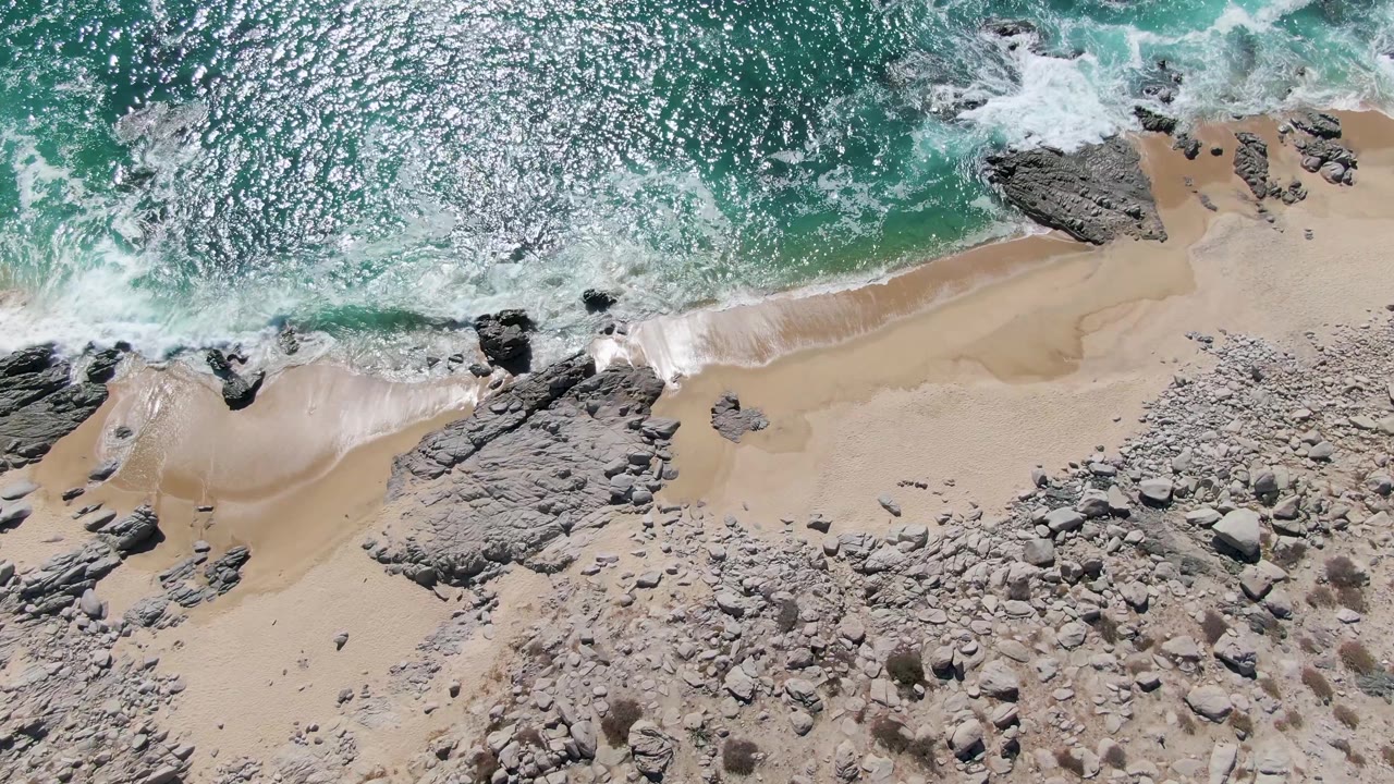 Overhead view at Buzzards Beach, Baja California Sur, with turquoise water, white surf, pale sand, gray rocks, and no visible people.