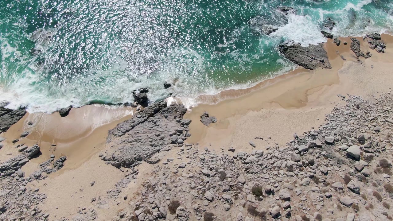 A bright overhead frame at Buzzards Beach shows sunlit turquoise water pushing foam along the sandy shoreline beside gray rock outcrops, with no visible people.
