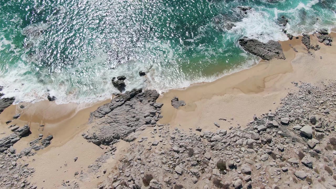 At Buzzards Beach, turquoise water fills the upper frame with strong sun shimmer while white water breaks onto sand and rocks below, with no visible people.