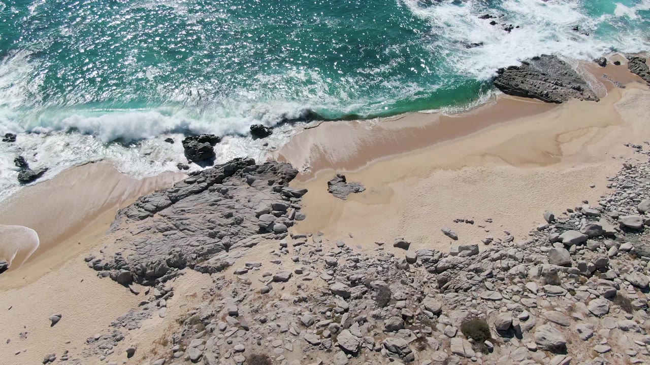 Overhead view of turquoise surf breaking onto sand and gray rock outcrops at Buzzards Beach, Baja California Sur, with no visible people.