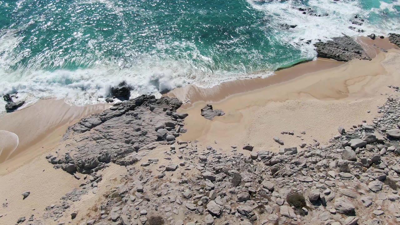 Aerial frame with white foam pushing farther onto the sandy shore beside a broad rock shelf at Buzzards Beach, Baja California Sur, with no visible people.