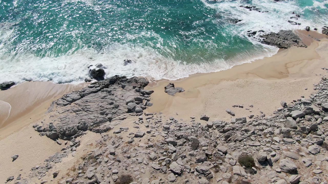 Foamy turquoise water curves around dark shoreline rocks and drains across pale sand at Buzzards Beach, Baja California Sur, with no visible people.
