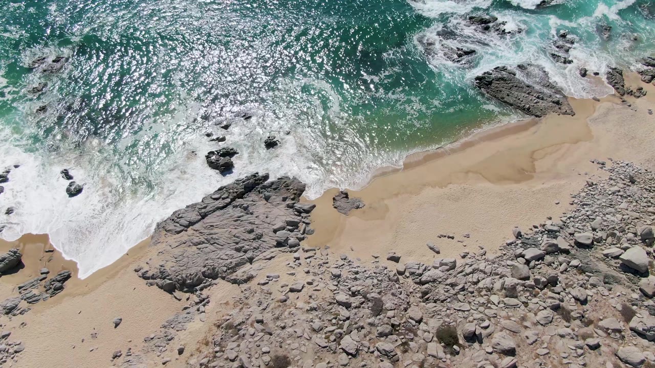 Aerial view of turquoise surf washing over sand and rocks.