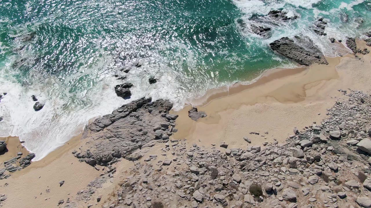 Aerial view at Buzzards Beach with white foam pulling back around scattered rocks, pale sand exposed below, turquoise water above, and no visible people.