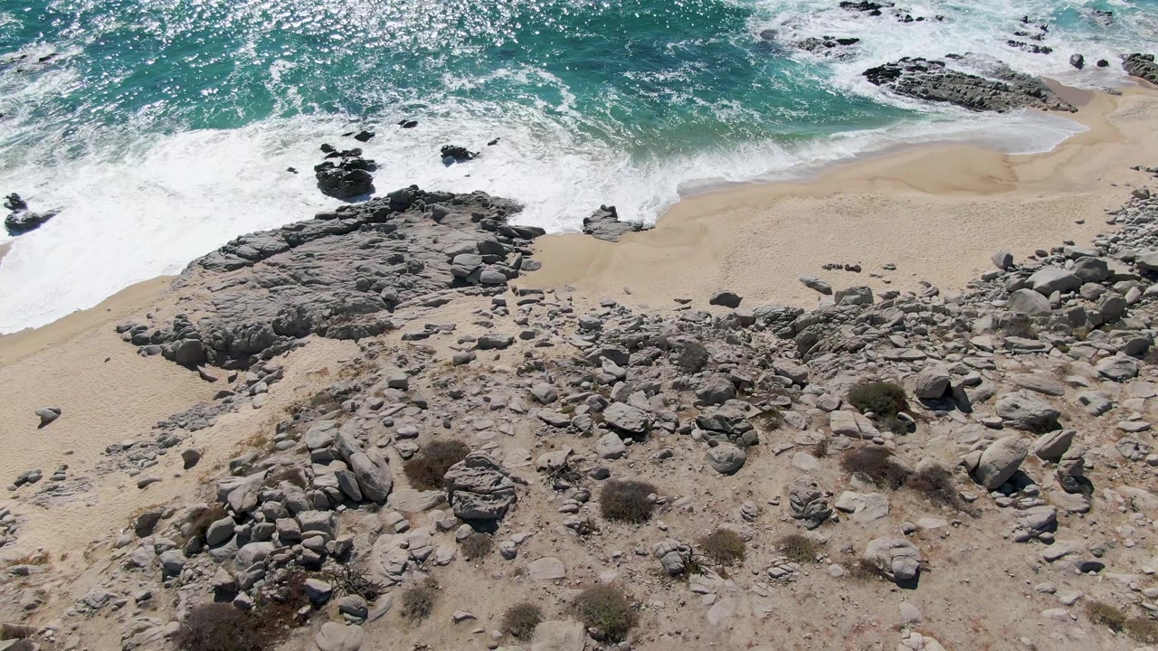 Aerial view of turquoise waves breaking onto a rocky sandy beach.
