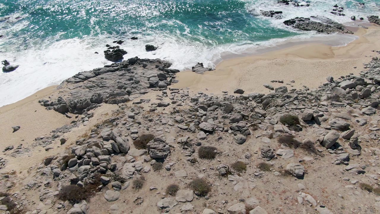 At 0:01, Buzzards Beach appears from above with foamy turquoise water spread farther along the sand, gray rock clusters below, and no visible people.