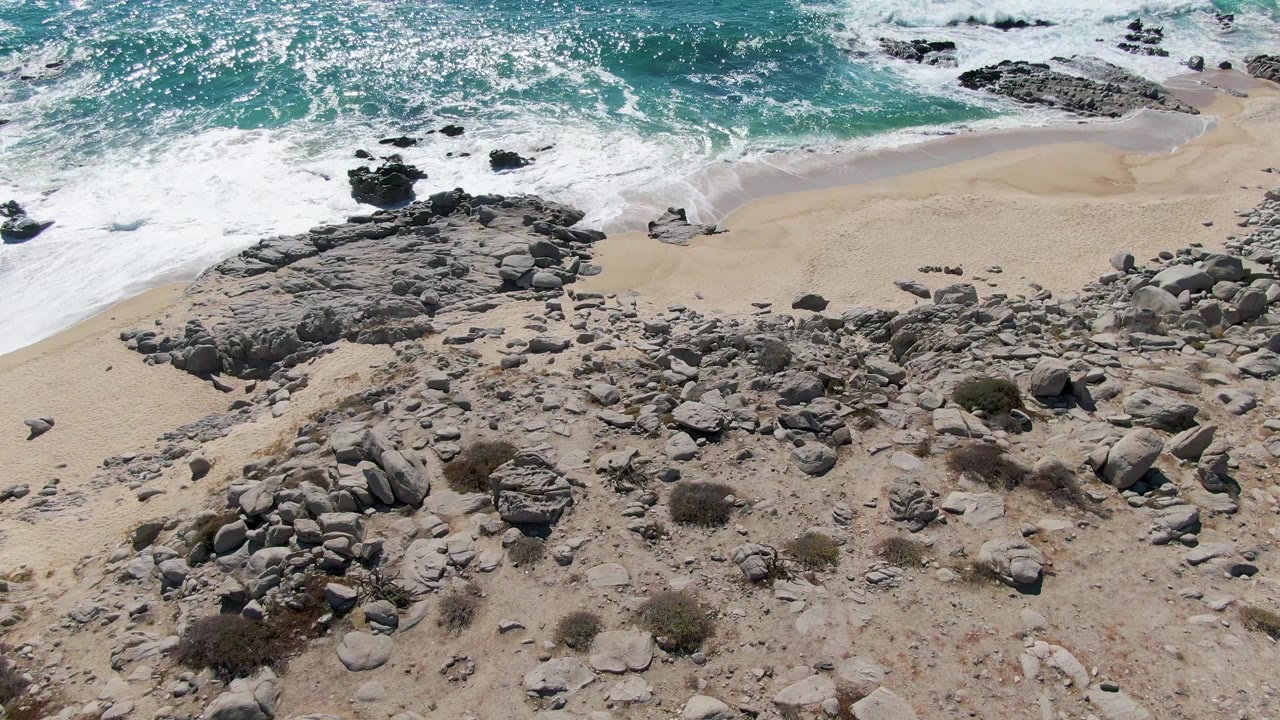 At 0:02, the aerial view holds on Buzzards Beach with breaking white water near the top edge, pale sand through the center, rocky ground below, and no visible people.