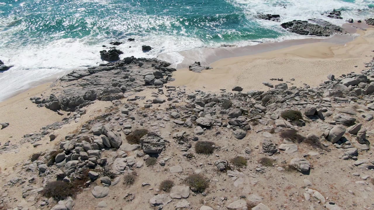 At 0:03, Buzzards Beach is seen from above as white foam curls onto sand beside dark shoreline rocks, with scattered stones and no visible people.